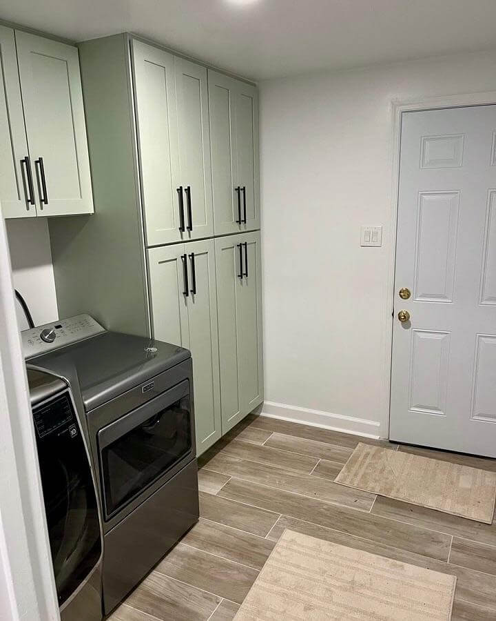Soft sage shaker cabinets with black handles and wood-look tile floor in laundry room from @blackstonehomeservices. Clean, modern green laundry room ideas.