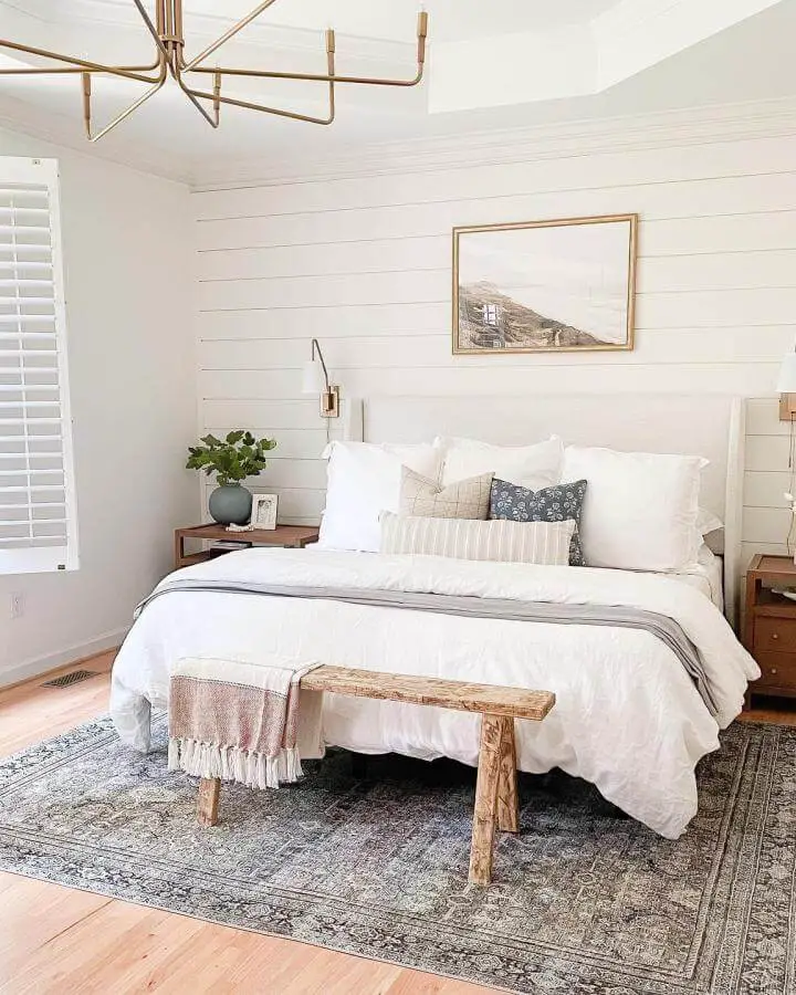 Serene white shiplap bedroom from @crawfordandcompany. Features brass chandelier and cozy neutral bedding with wooden bench.