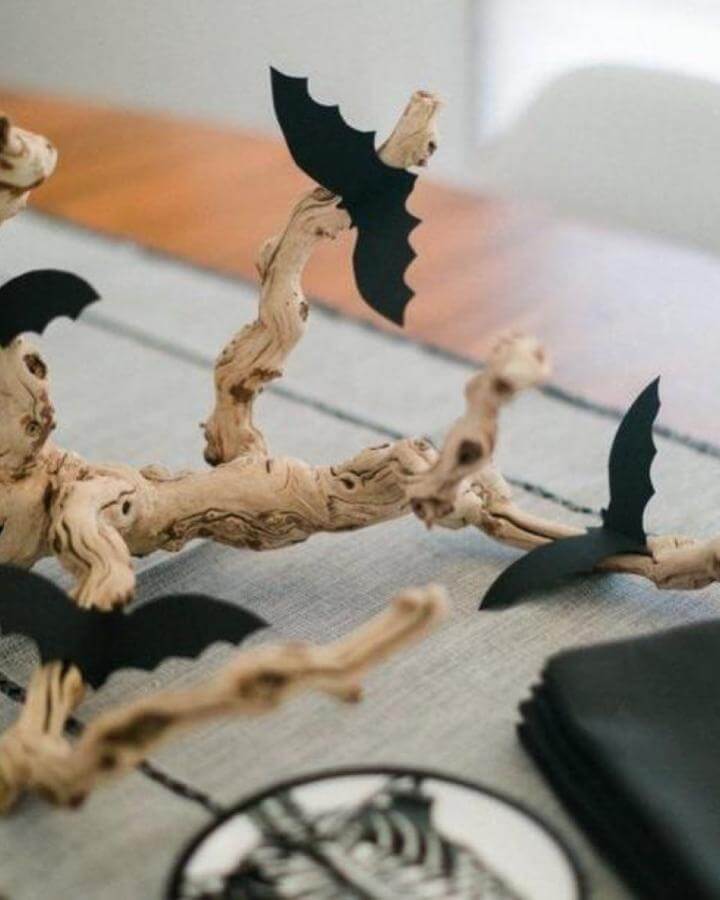 Weathered driftwood branch centerpiece from @craftgawker featuring black paper bat cutouts clipped along twisted natural wood branches.