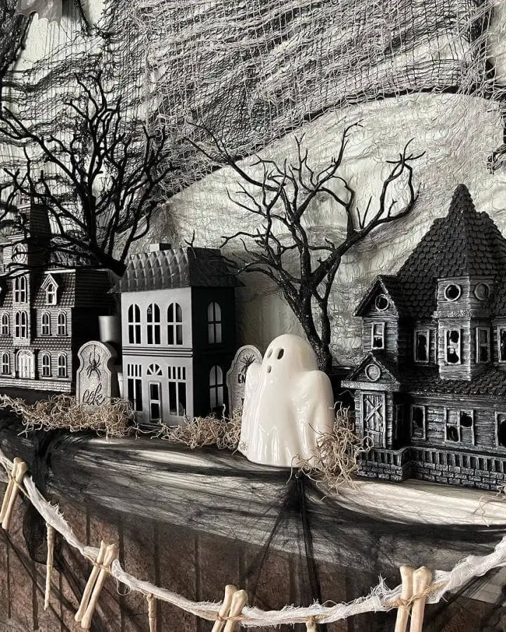 Monochromatic Halloween village display from @designed_comfort_4_5. Ghost figure positioned among miniature houses and bare branches on wooden platform.