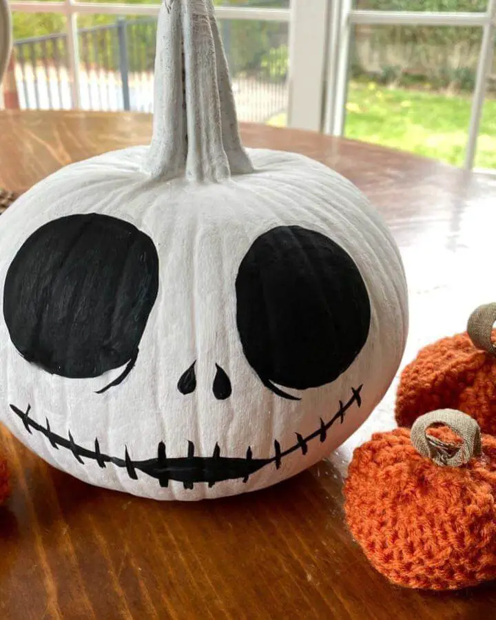 White painted pumpkin with black eyes and stitched mouth from @exquisitelyunremarkable. Orange knitted pumpkins nearby.