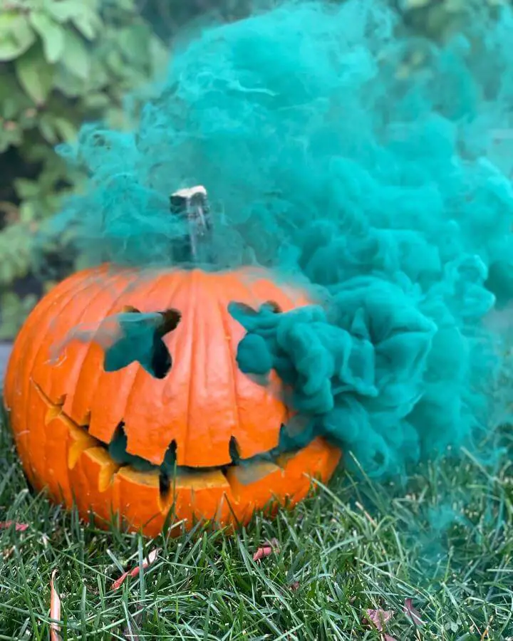 Bright orange carved pumpkin from @peacocksmoke surrounded by vibrant turquoise smoke effects on grass.