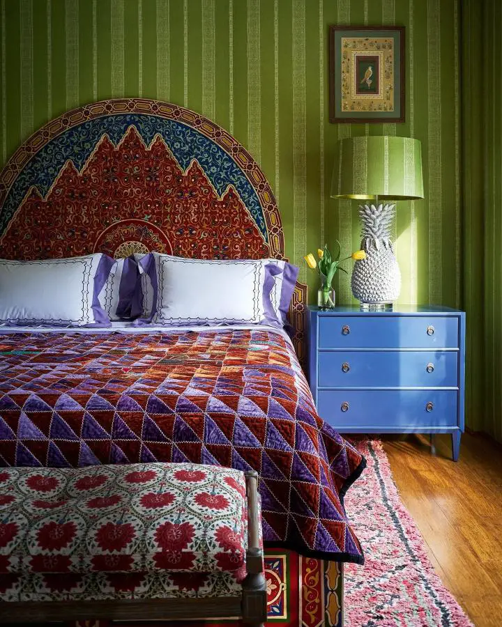 Ornate Moroccan headboard bedroom from @homesandgardensofficial featuring rich red and blue patterns against green striped walls.