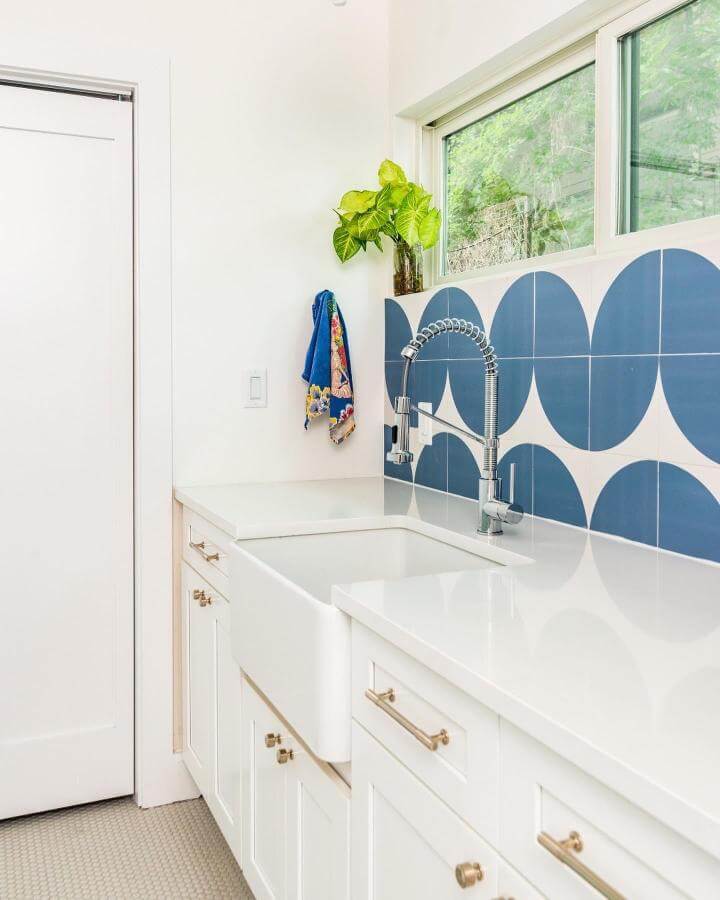 White farmhouse sink with blue geometric tile backsplash and brass hardware in a laundry space by @ruvati. Bright and modern with a clean design.