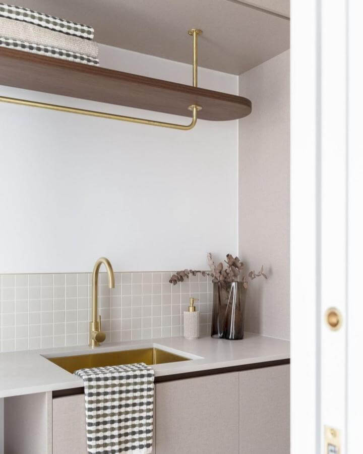 Brass faucet and sink in beige tiled laundry nook with wooden shelf from @queenslandhomes. Modern warmth and style combined.