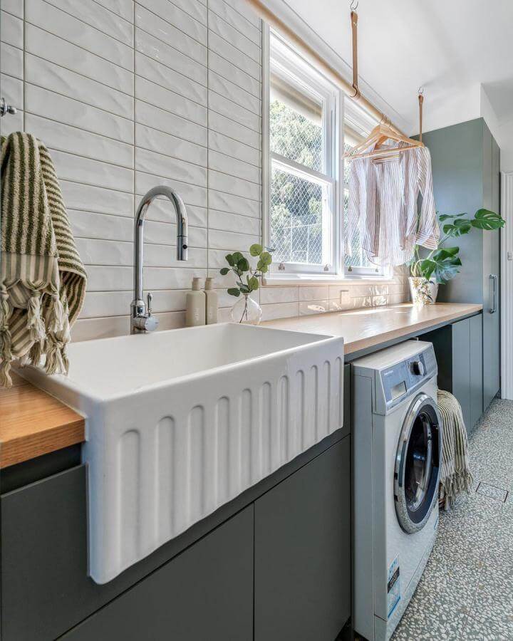 White ceramic farmhouse sink with ribbed design and chrome faucet in a laundry room by @fienza.au with wood countertop and green cabinetry.