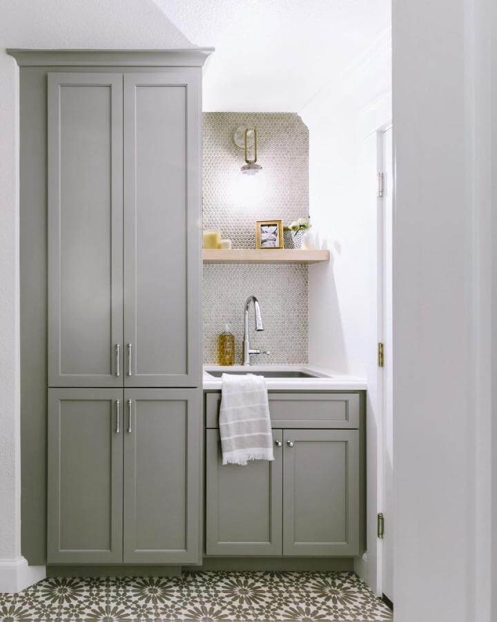 Gray shaker cabinets and pebble tile backsplash laundry space from @woodcliffbuilders featuring brass hardware and patterned floor tiles.