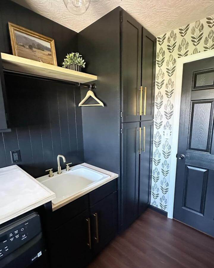Black matte cabinetry laundry room with brass handles and wooden shelf by @bookgirlcreations featuring high contrast wallpaper and modern sink setup.