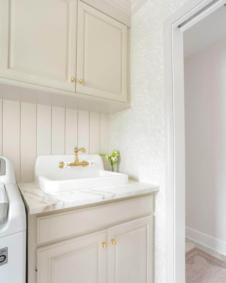 Creamy farmhouse sink with brass fixtures in a classic laundry space by @ducydesign. Marble countertop and paneled backsplash detail.
