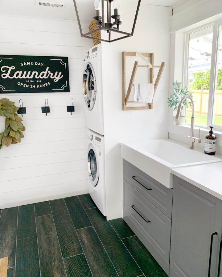 White farmhouse sink with gray cabinetry laundry room design from @dreamingofhomemaking featuring stacked washer dryer and wood-look tile floor.