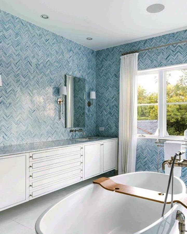 Light blue chevron patterned tile wall in bathroom from @meredithsteinhartdesign with white tub and brass fixtures creating calm light blue bathroom ideas ambiance