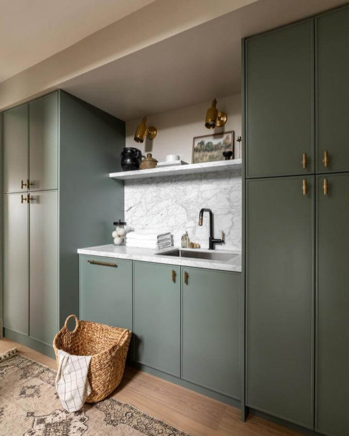 Soft sage green cabinetry and marble countertop laundry room from @maria.decotiis. Elegant brass fixtures and natural woven basket enhance luxury laundry room ideas.