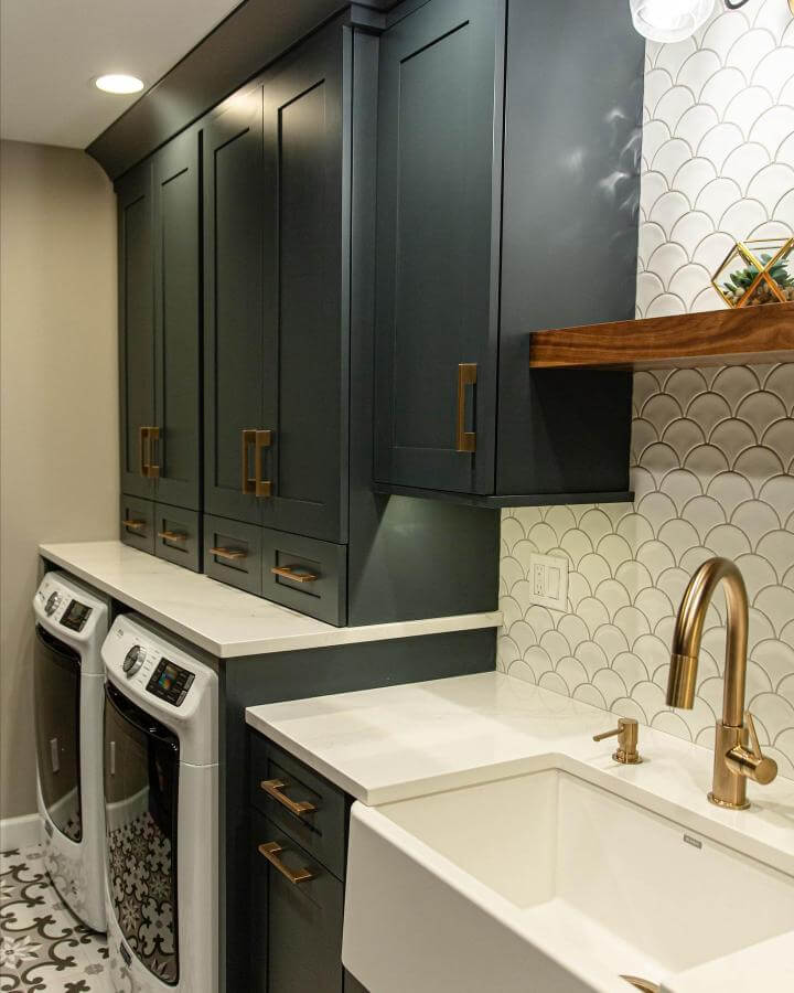 Dark navy cabinets with brass fixtures and scalloped tile backsplash in a luxury laundry room by @bellacasa_kb, showing quartz countertops and a farmhouse sink.