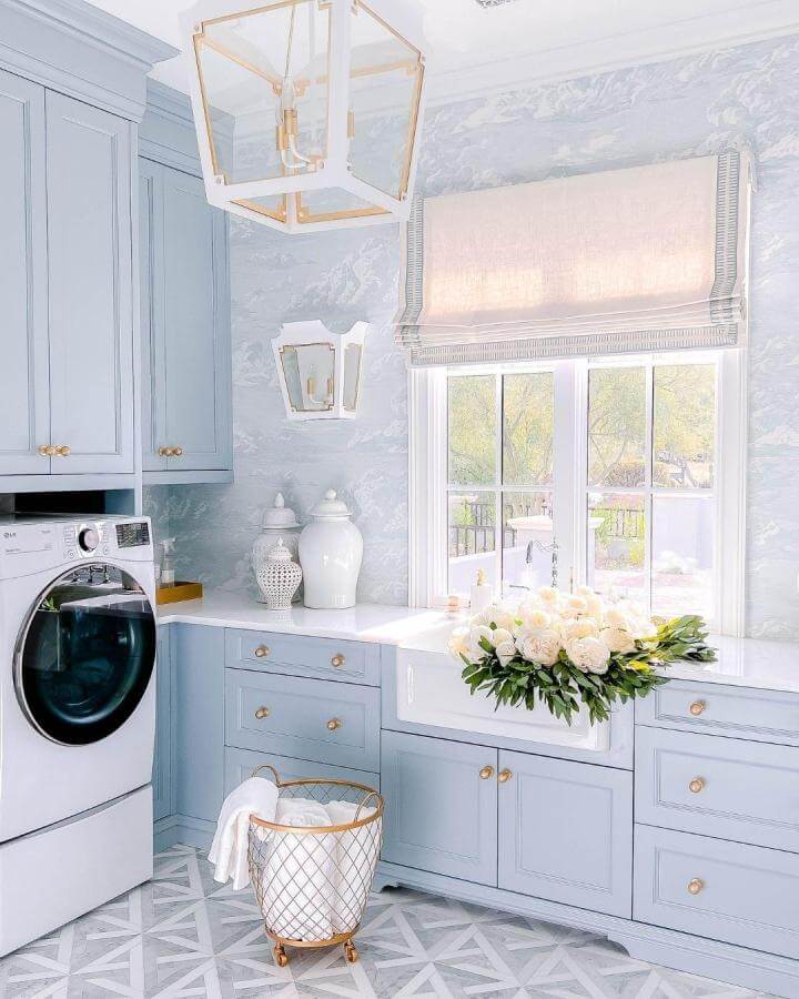 Soft blue cabinetry with gold hardware and geometric floors in a bright luxury laundry room by @armacmartin featuring marble counters and floral decor.