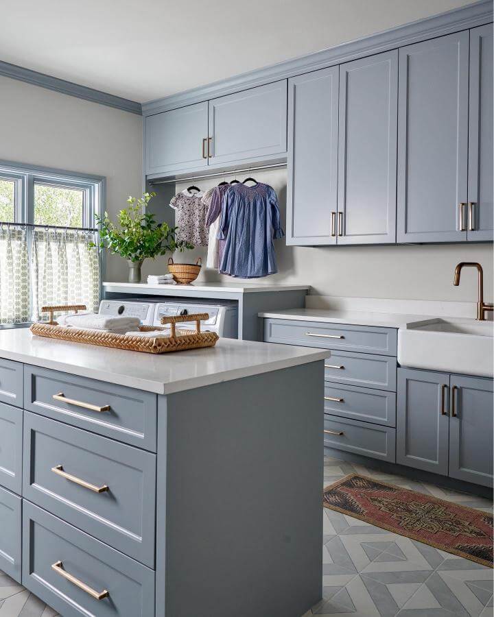 Soft blue cabinetry and quartz island in a luxury laundry room ideas space by @morganfarrowinteriors with farmhouse sink and bronze faucet.
