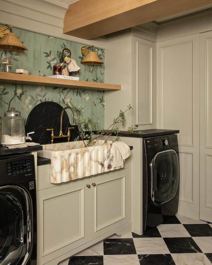 Creamy veined marble farmhouse sink with black and gold fittings in a sage green floral wallpaper luxury laundry room by @koehndesign_livebeautiful. Elegant and chic space with checkered floor.