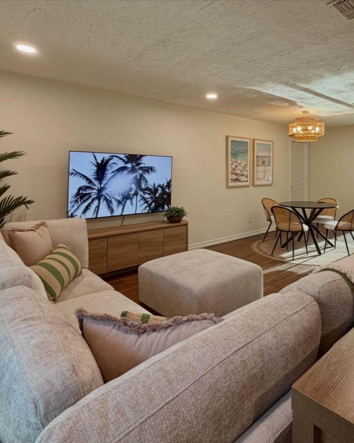 Beige sectional sofa with green striped pillows and natural rattan chairs in a warm coastal-inspired living and dining room from @stayatpalmandtide featuring woven textures and soft wood tones.