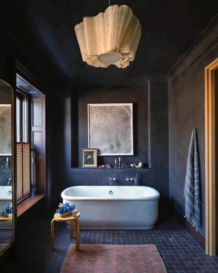 Charcoal dark walls bathroom with white soaking tub and wooden accents from @trevortondro. Cozy and textured low-light interior.