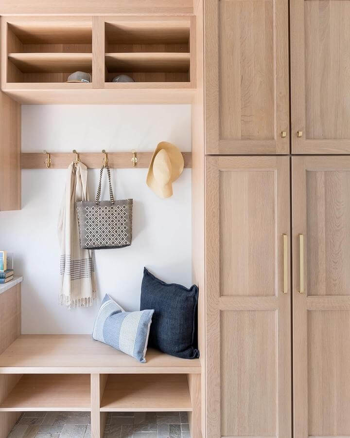 Light oak wood mudroom bench with brass hooks and navy blue pillows from @grayoakstudio. Cozy and organized entry space.