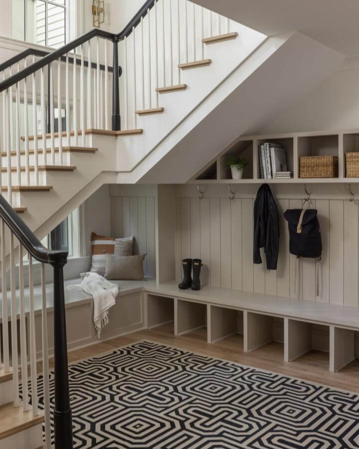 Light gray wooden built in bench and cubby storage with hooks and cozy seating nook under stairs by @wellhouseandco. Geometric black and white rug complements the design.