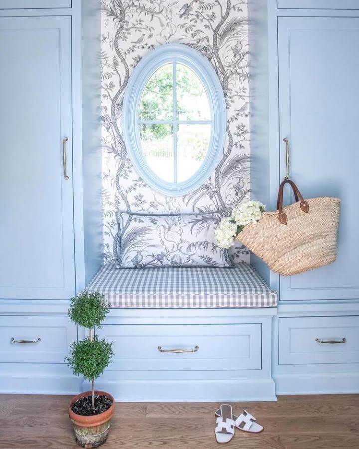 Light blue built in bench with gingham cushion and floral wallpaper behind oval window in mudroom by @clarybosbyshell.