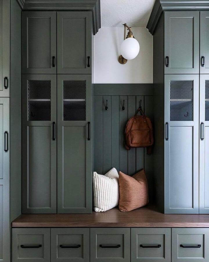 Dark green built-in cabinets and wooden bench with pillows in a stylish entryway from @saralynnbrennan featuring black hardware and mesh storage panels.