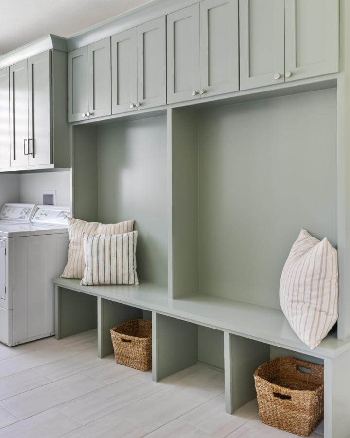 Soft sage green built-in mudroom bench with wicker baskets and striped pillows from @structurdesign. Functional and welcoming entryway seating.