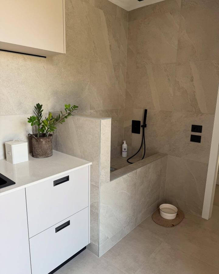 Beige stone tiled dog wash area with matte black fixtures and simple plant accents by @pippotheweim in a modern mudroom setup.
