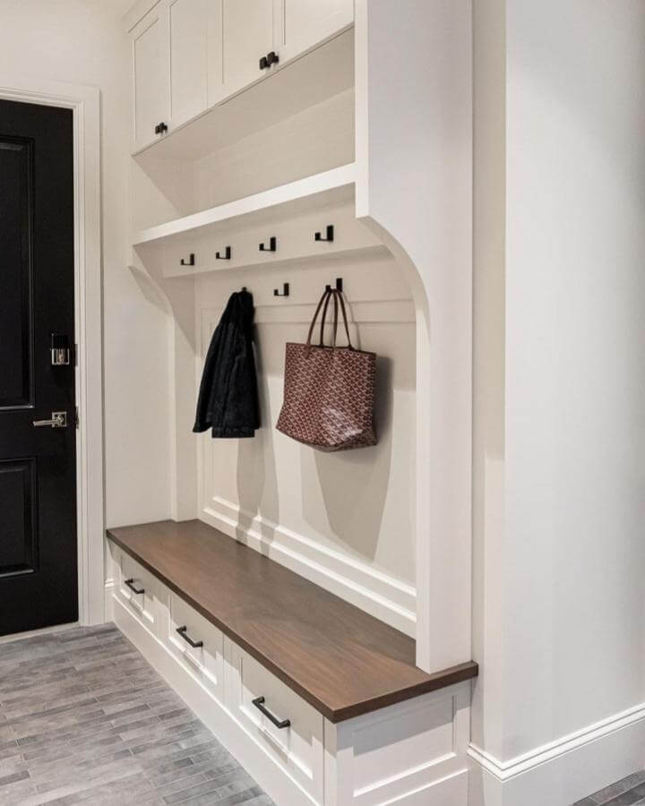 Lacquered white cabinetry with wooden bench and black hooks from @verandaestatehomes showcasing mudroom shoe storage ideas with drawers and hanging space.