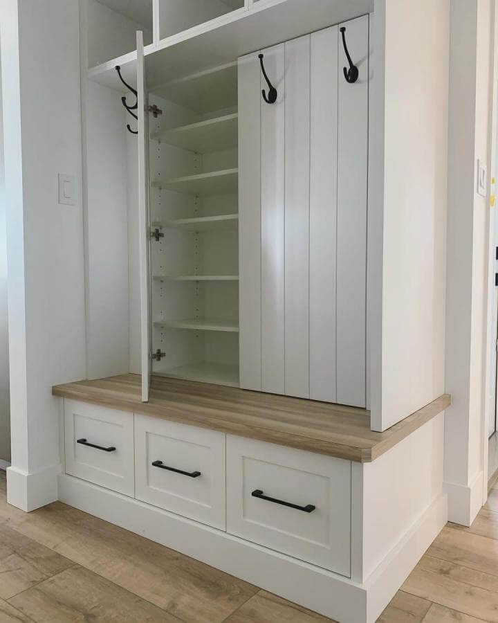 White built-in storage bench with wood top and black handles in mudroom by @wall.to.wall.design Featuring vertical shoe shelves and hanging hooks.
