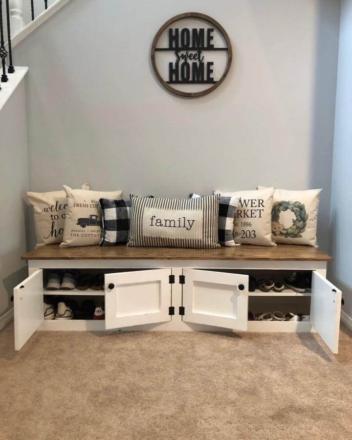 White wooden bench with brown top and decorative pillows showing concealed shoe storage under seating from @rustiquebyjandp in cozy mudroom corner.