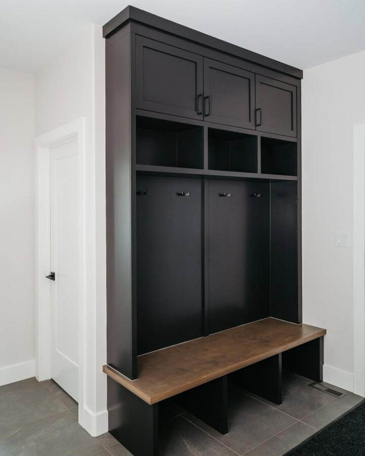 Black built-in mudroom bench with wooden seat and open cubbies from @jocelynbydesign. Functional and stylish shoe storage with hooks and compartments.
