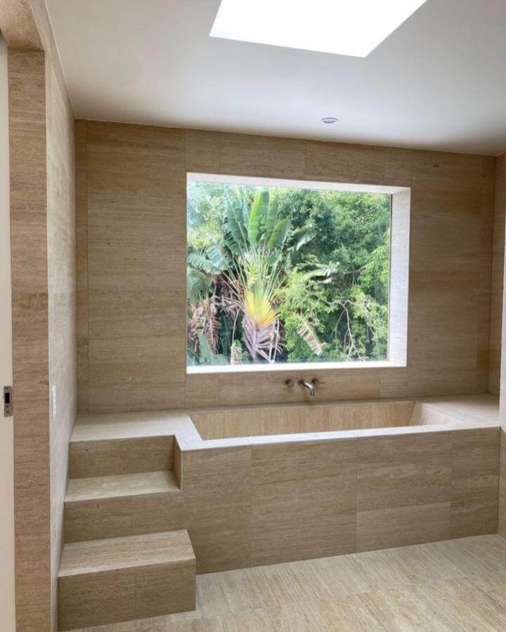 Creamy beige travertine bathtub and walls from @ceramicasenio natural stone bathroom ideas with large window and greenery outside.