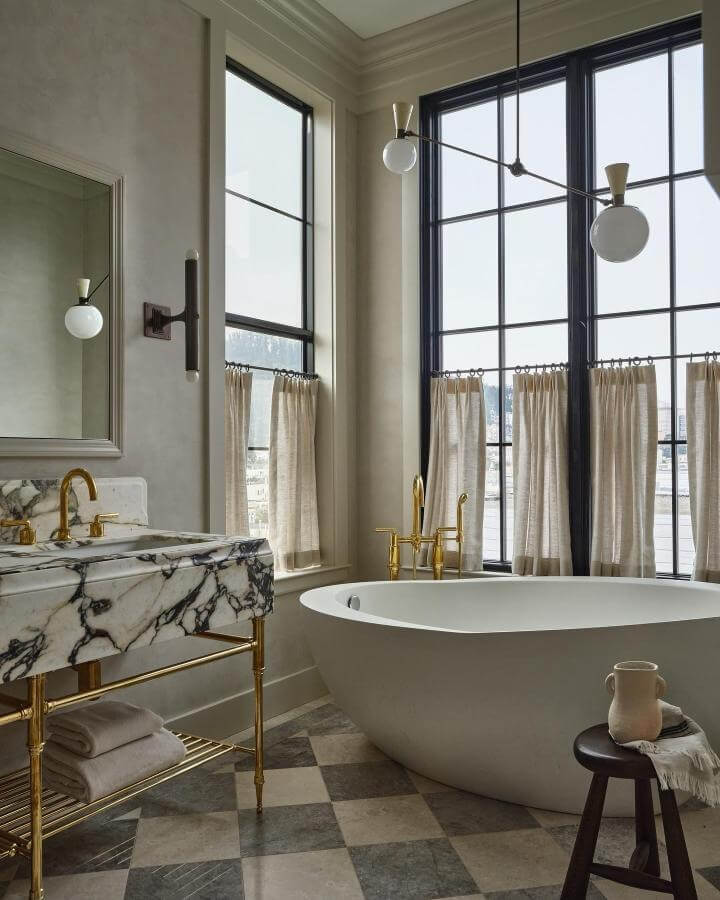 White marble vanity with dark veins and brass fixtures in a bright bathroom by @michaelcliffordphotography. Natural stone bathroom ideas with checkered stone floor and tall windows.