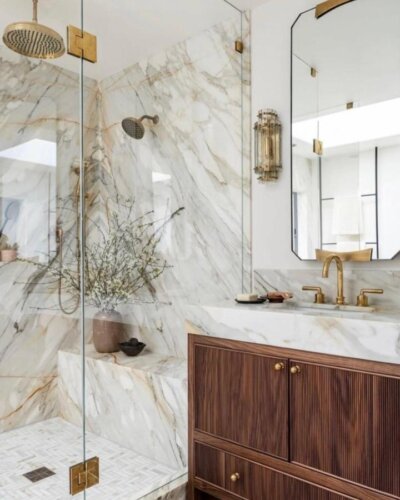 Creamy white marble shower with brass fixtures and wooden vanity from @caskro. Elegant natural stone bathroom ideas with warm accents.