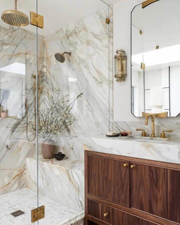 Creamy white marble shower with brass fixtures and wooden vanity from @caskro. Elegant natural stone bathroom ideas with warm accents.