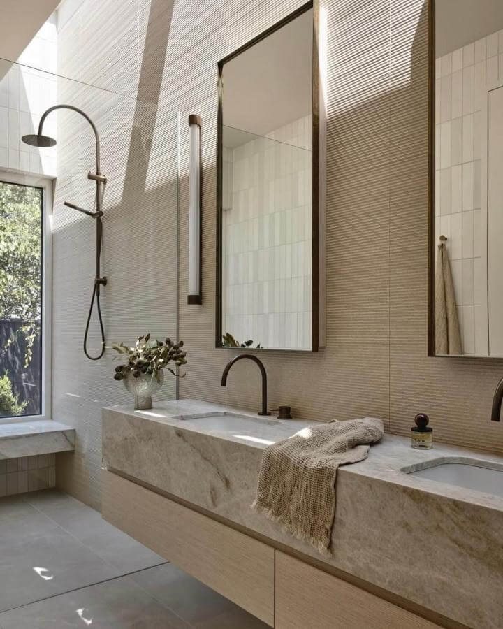 Beige textured tile walls and smooth marble countertop bathroom vanity from @derek_swalwell natural stone bathroom ideas with bronze fixtures and soft natural light.