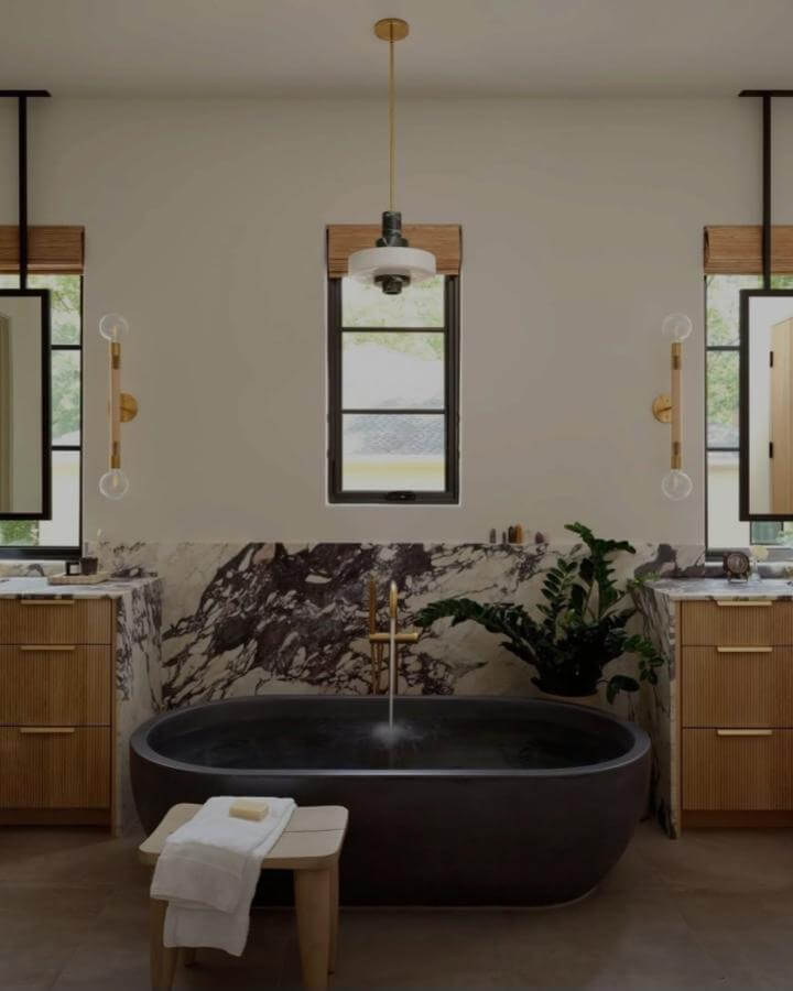 Dark marble accent wall with matte black stone freestanding tub and wood cabinetry from @seanlitchfield natural stone bathroom ideas with plant accents and warm lighting