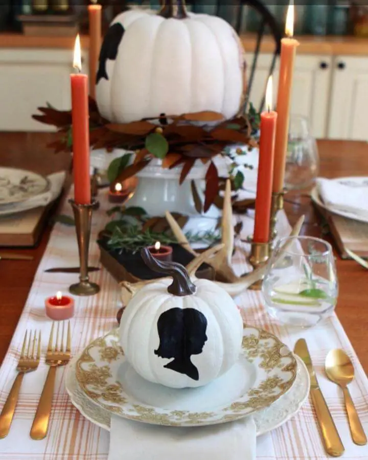 White pumpkin with black silhouette design from @baker_nest. Elegant fall tablescape with candles and autumn leaves.