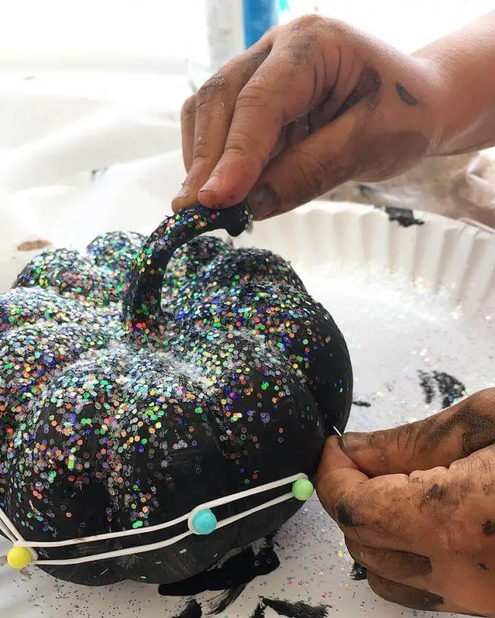 Sparkly black pumpkin craft from @k.e.artforkids. Hands applying holographic glitter to create festive Halloween decorations.