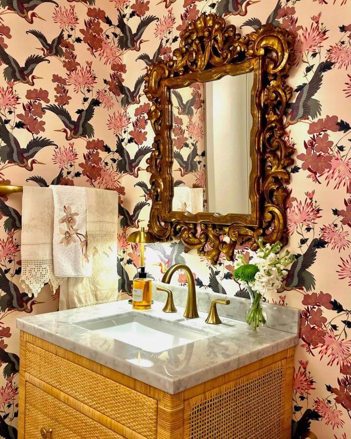 Vintage gold ornate mirror above marble and rattan vanity in pink powder room ideas with floral crane wallpaper and brass fixtures from @katewalker_design. Charming and elegant style.