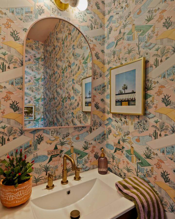 Pink wallpaper with vintage motel scenes and brass fixtures in a powder room from @schumacher_southafrica. Cozy and playful pink powder room ideas.