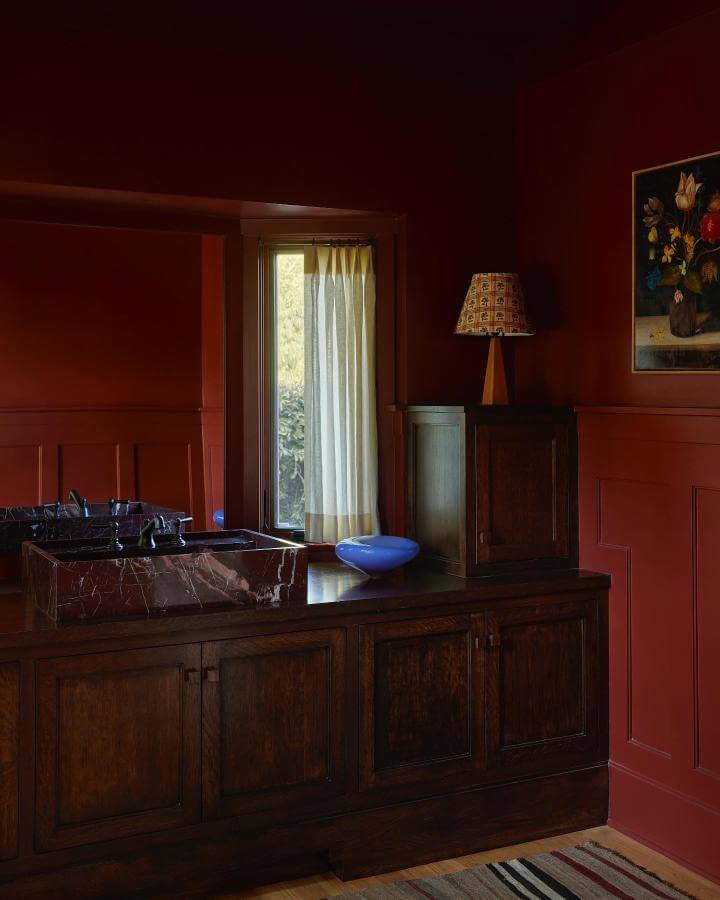 Deep red walls with rich wood cabinetry and marble sink in a moody red powder room ideas setting by @electricbowery, classic lamp and artwork included.