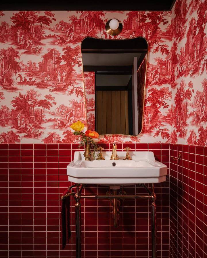 Red toile wallpaper and glossy red subway tiles in a powder room with brass fixtures and a white porcelain sink from @archdigest. Bold red powder room ideas.