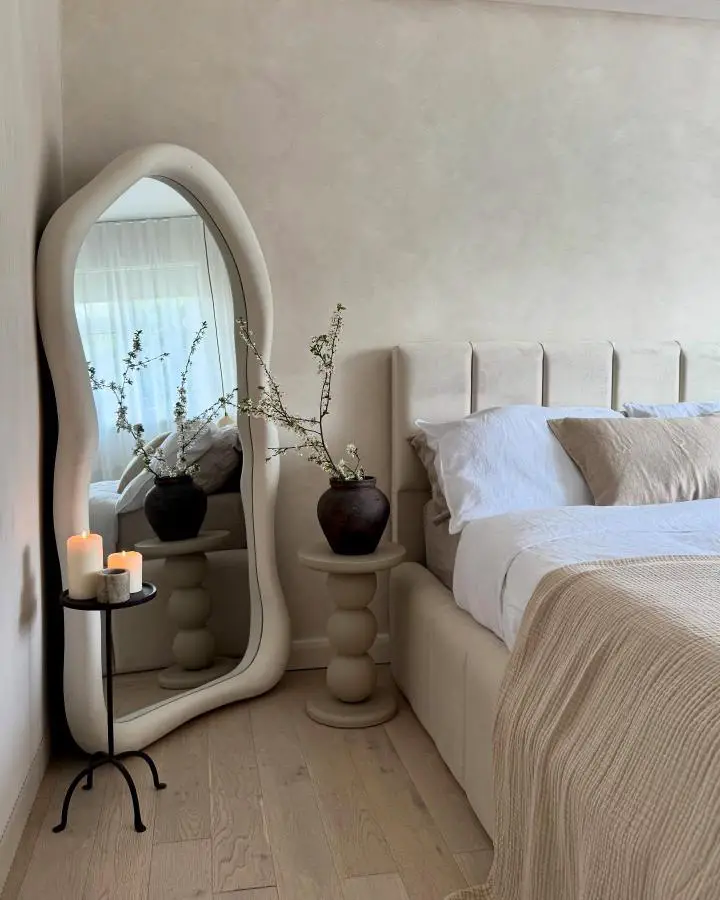 Curved beige mirror from @home_of_harris. Sculptural nightstand with candles and neutral bedding create calming atmosphere.