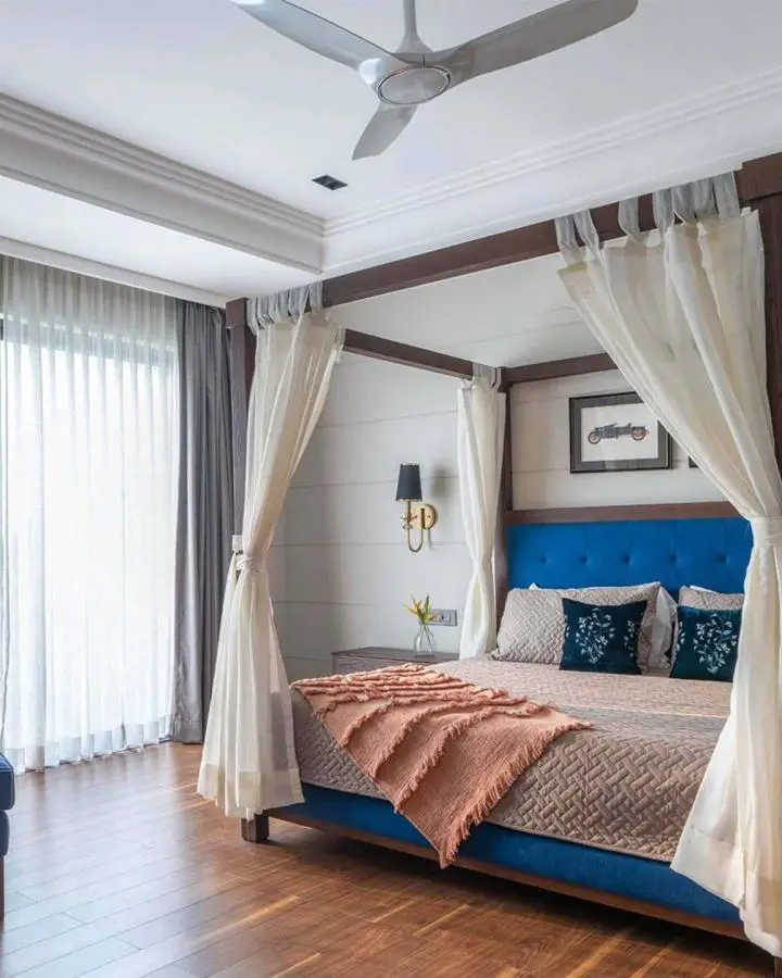 Blue upholstered canopy bed from @spdc_studio. Cream curtains frame the wooden four-poster design with coral throw blanket.