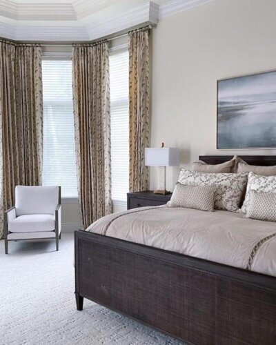 Neutral bedroom with corner windows and patterned curtains from @sandraasdourianinteriors ⁠. Dark wood furniture complements beige color scheme.