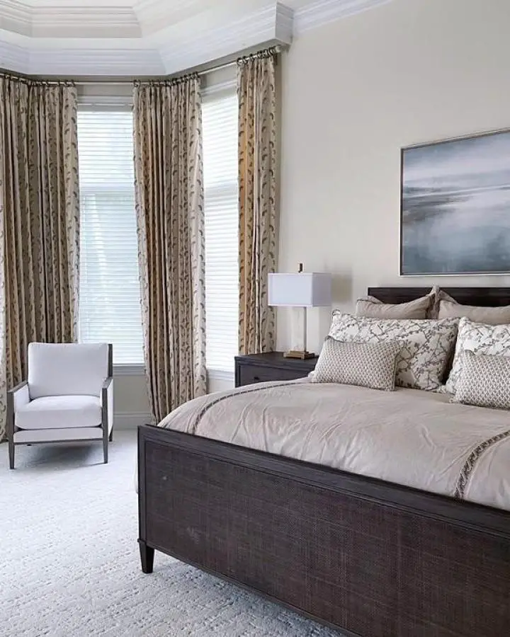 Neutral bedroom with corner windows and patterned curtains from @sandraasdourianinteriors . Dark wood furniture complements beige color scheme.