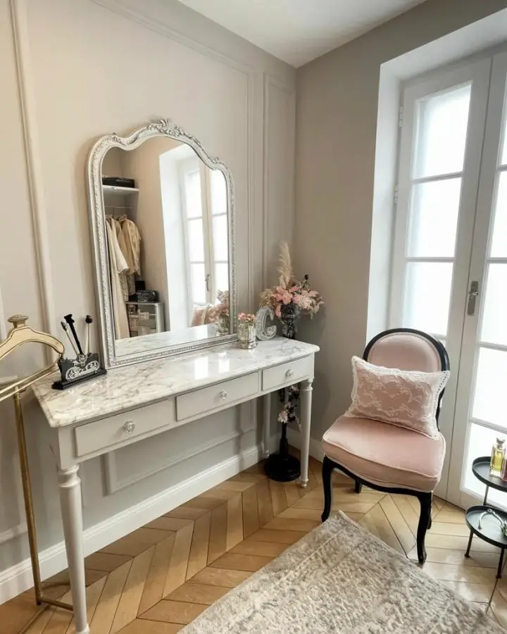 Ornate silver-framed mirror above white marble vanity table with pink upholstered chair and natural lighting.