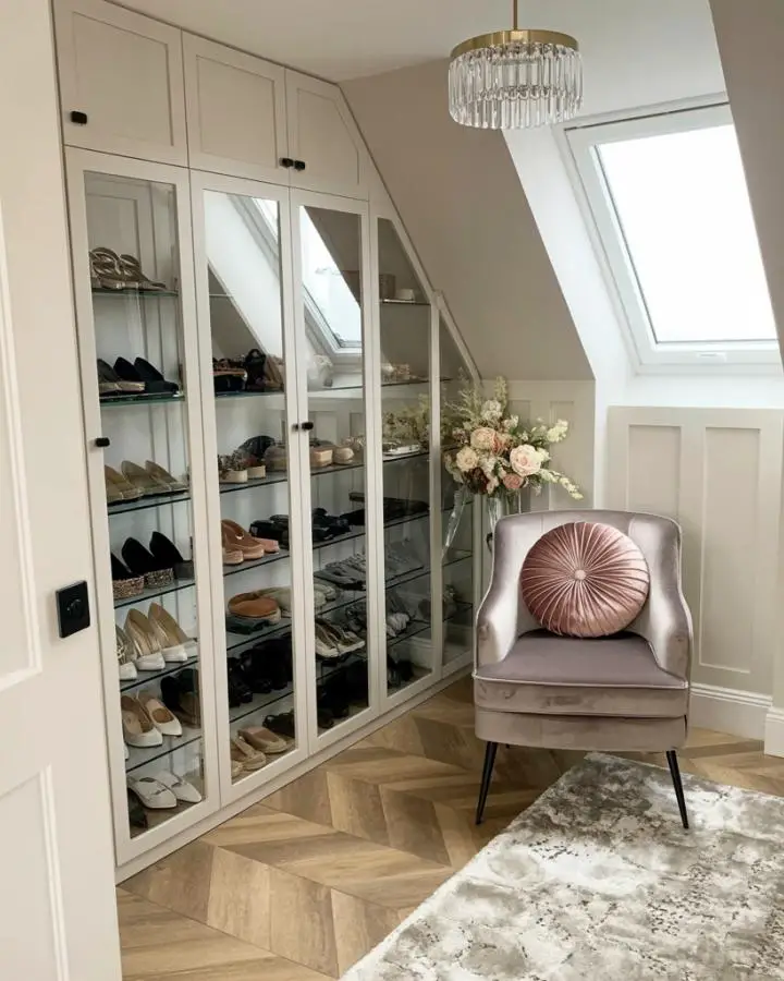 White glass-front cabinet system displaying organized shoe collection in compact attic space with natural lighting.
